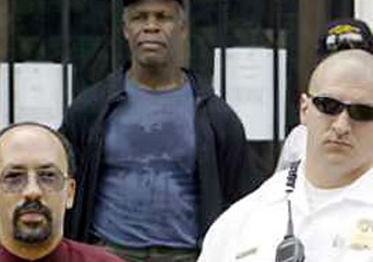 Three men stand together in a 2004 photograph of Danny Glover being arrested. They are positioned in a group composition with one person standing slightly behind two others. The individual in the center wears a navy blue t-shirt and dark jacket, while the person on the left wears a maroon colored shirt, and the person on the right wears a white shirt and sunglasses. The background shows what appears to be a building entrance with glass doors or windows.