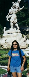 Vintage 2004 travel photograph showing a tourist posing in front of a classical stone statue monument. The subject wears a casual blue t-shirt and shorts on a sunny day, demonstrating classic pre-social media era vacation photography. This authentic throwback travel snapshot represents early-2000s tourism documentation, capturing the traditional 'I was here' moment commonly found in personal photo albums before the advent of Instagram and selfie culture. The composition features the full statue in the background while the tourist stands centered in the foreground, exemplifying typical point-and-shoot camera vacation photography of the period.